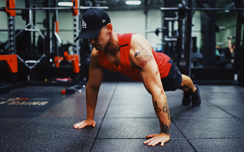 Lean muscular man wearing a red tank top doing a plank in the gym